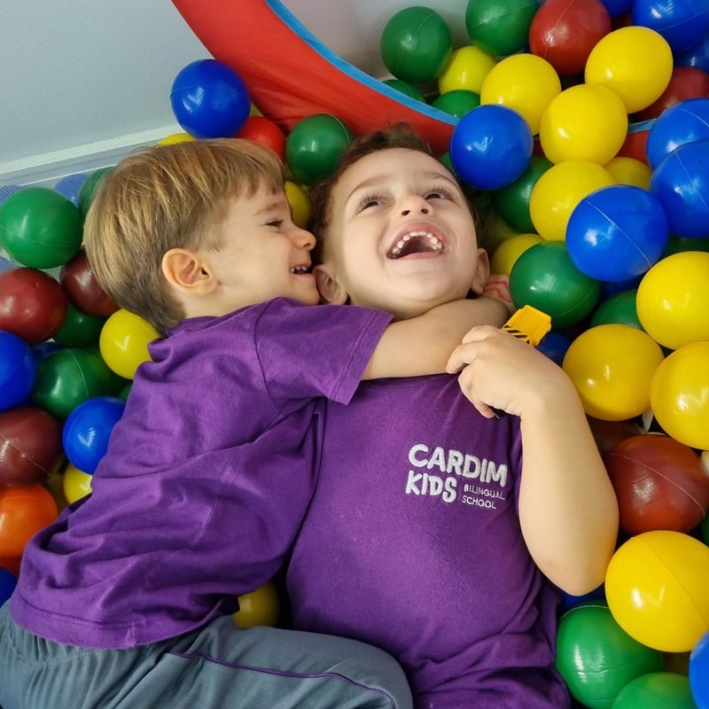 Duas crianças da Cardim Kids abraçadas e sorrindo dentro da piscina de bolinhas, vestindo o uniforme roxo da escola
